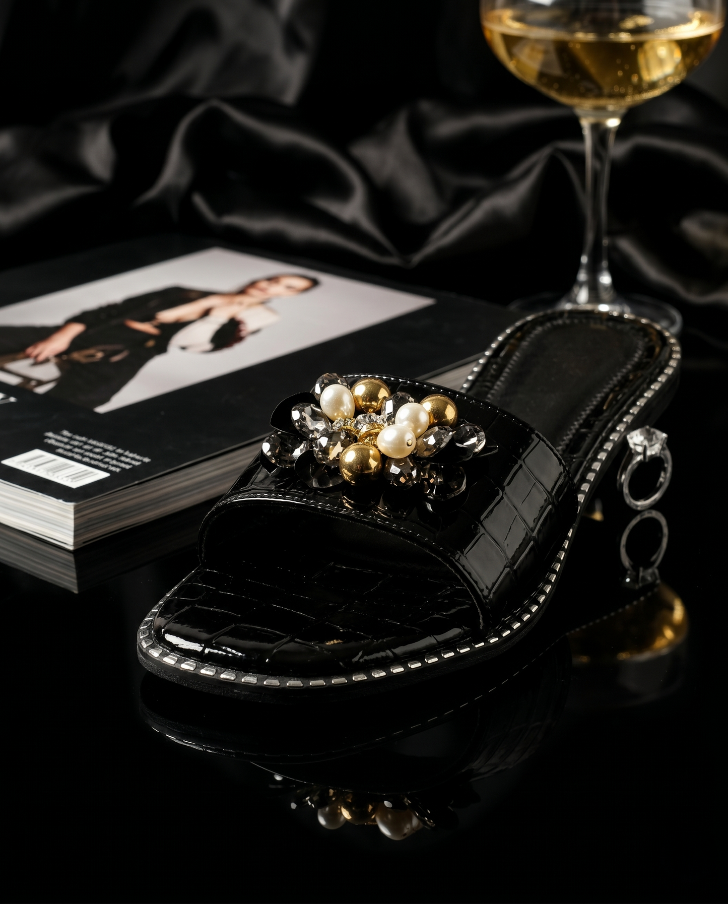 Topaz Black Flat Sliders on black surface with champagne coupe and magazine — black patent croc sliders with gold bead and pearl floral brooch embellishment by Solemates by Palak