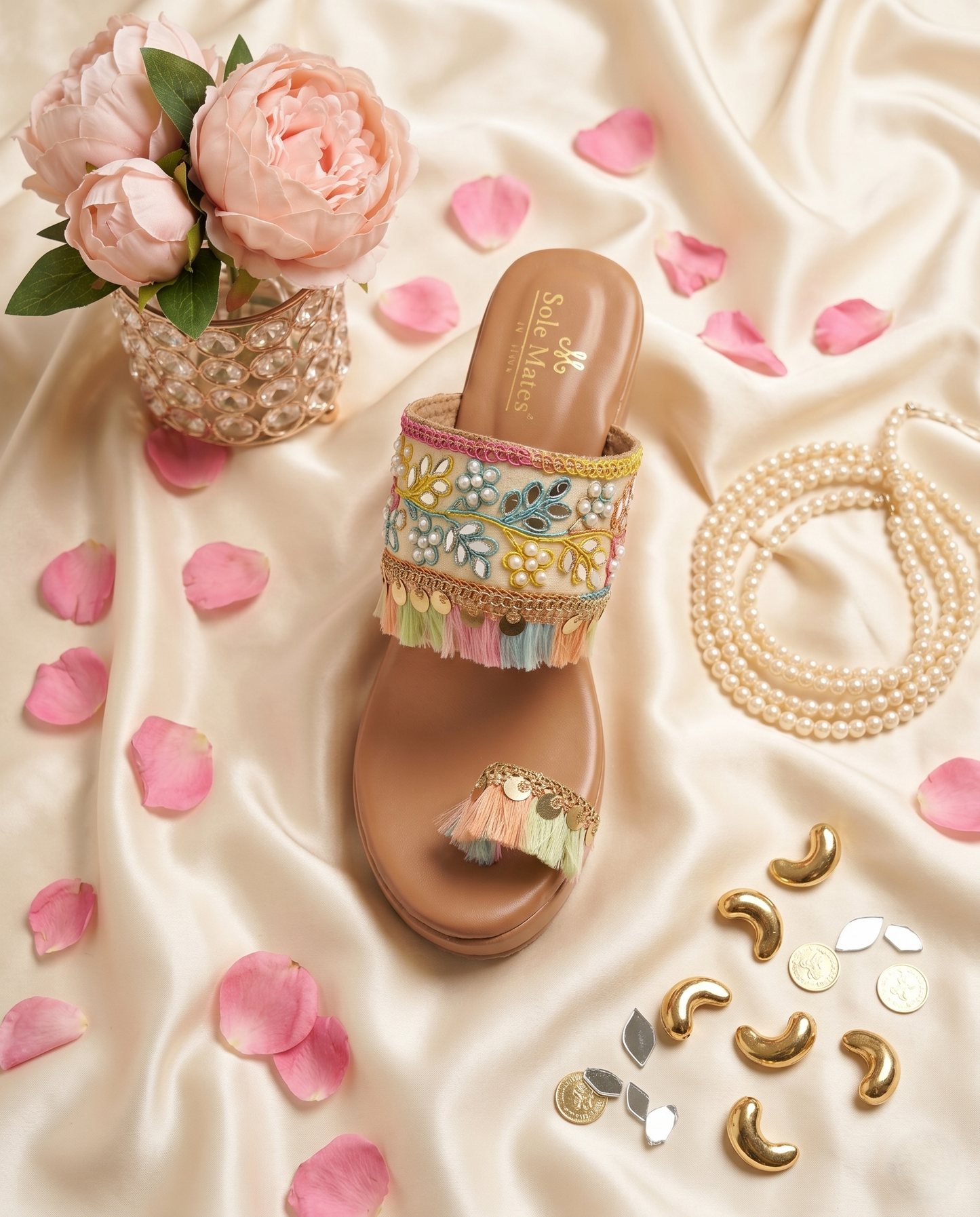 Top view of Gypsey Kohla Heels by Solemates by Palak on cream satin — wide embroidered strap with multicolour threadwork, mirror work, pearls and gold coin tassel fringe, pink peonies backdrop