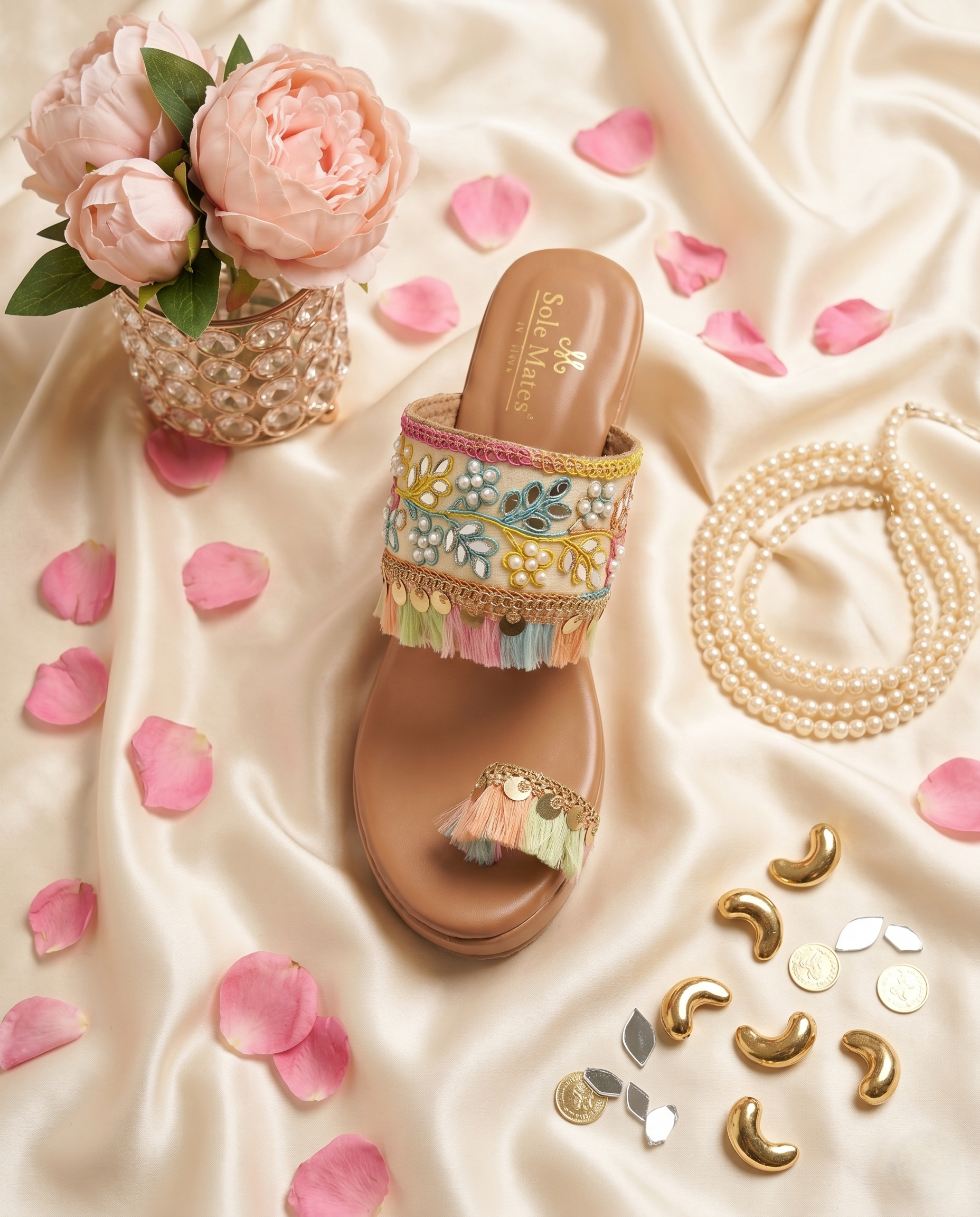 Top view of Gypsey Kohla Heels by Solemates by Palak on cream satin — wide embroidered strap with multicolour threadwork, mirror work, pearls and gold coin tassel fringe, pink peonies backdrop
